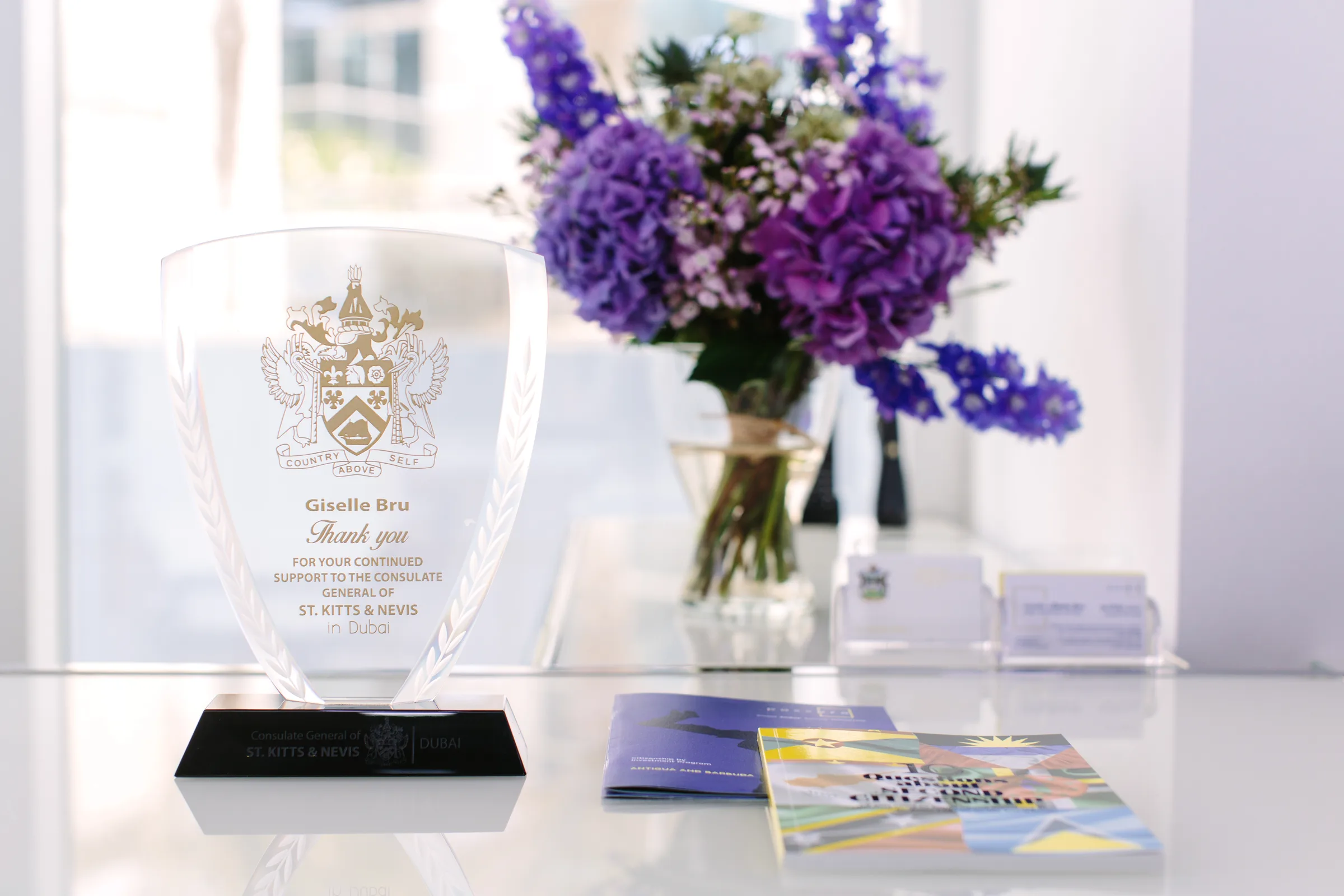 The Consulate General of St. Kitts and Nevis (Dubai) engraved-glass award presented to Giselle Bru, alongside PassPro programme brochures and delphinium florals.